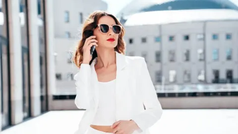 Poslovna žena u bijelom odijelu s naočalama razgovara na mobitel ispred zgrade