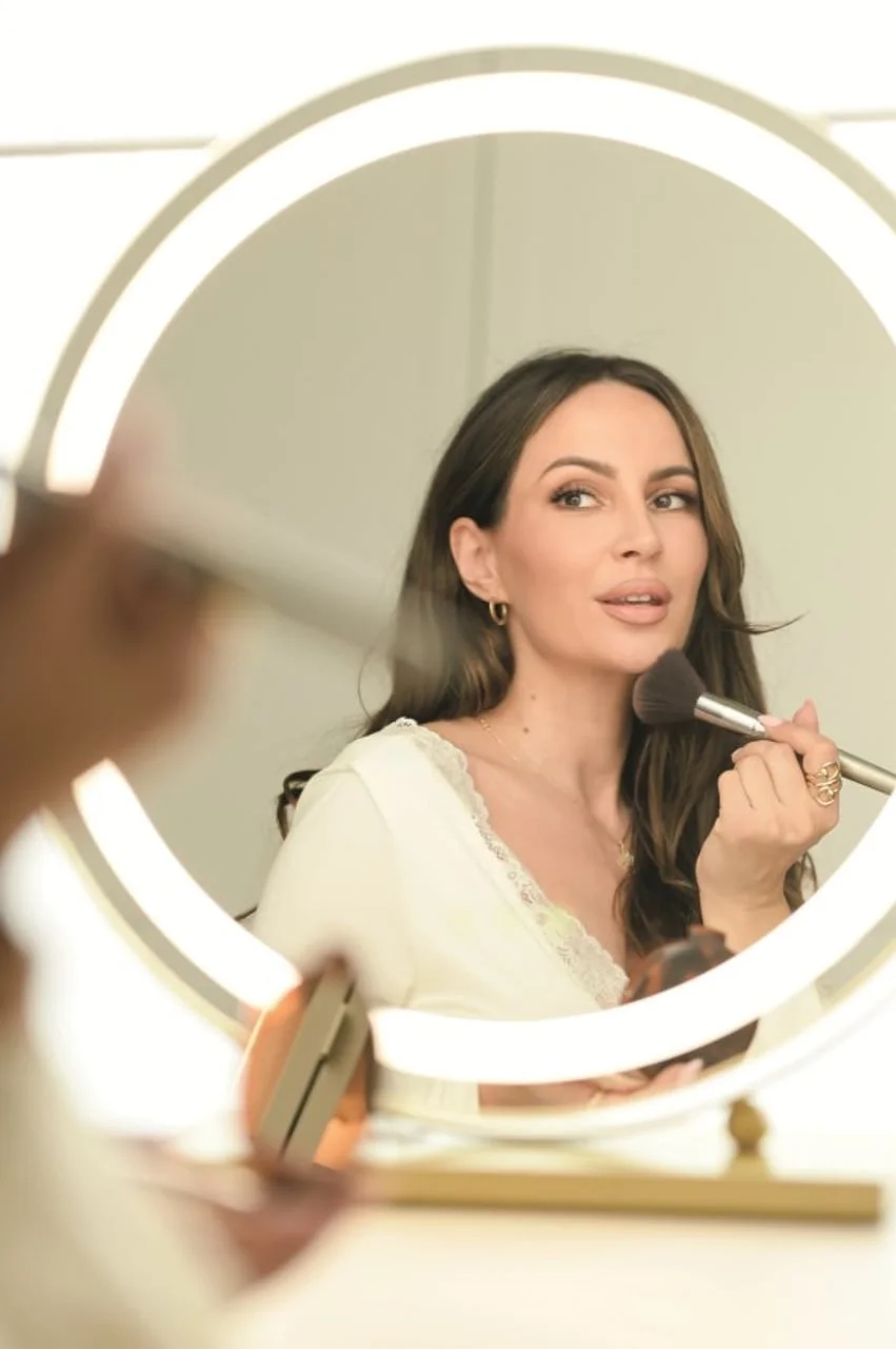 Odraz žene u okruglom ogledalu s rasvjetom dok nanosi šminku kistom.