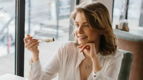 Mlada žena u bijeloj košulji smiješi se dok jede desert u restoranu.