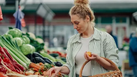 Žena na tržnici pažljivo bira svježe povrće uključujući mrkvu i papriku.