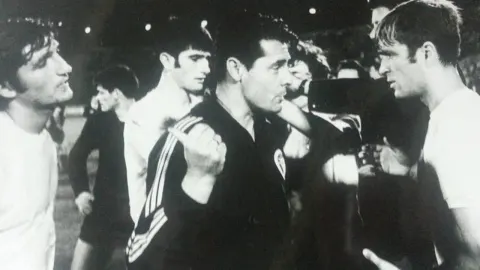 Crno-bijela fotografija Dušana Nenkovića u razgovoru s nogometašima Hajduka na terenu.