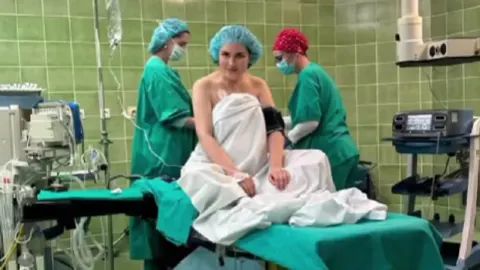 Lana Jurčević sjedi na bolničkom krevetu u operacijskoj sali okružena medicinskim osobljem.