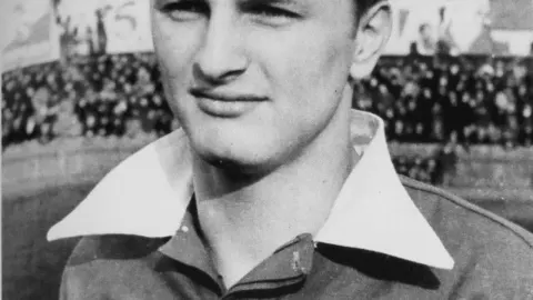 Crno-bijeli portret mladog nogometaša u dresu s bijelom kragnom na stadionu.