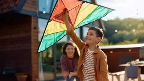 Dječak drži šarenog zmaja u zraku ispred kuće dok ga majka promatra.