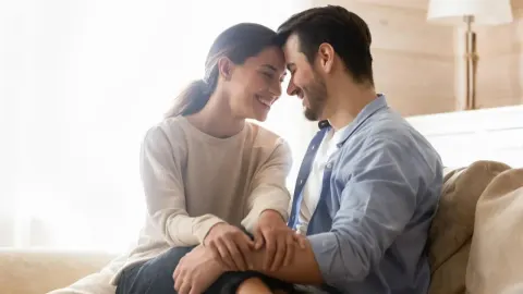 Nasmijani par sjedi blizu na kauču i dodiruje se čelima u dnevnom boravku.