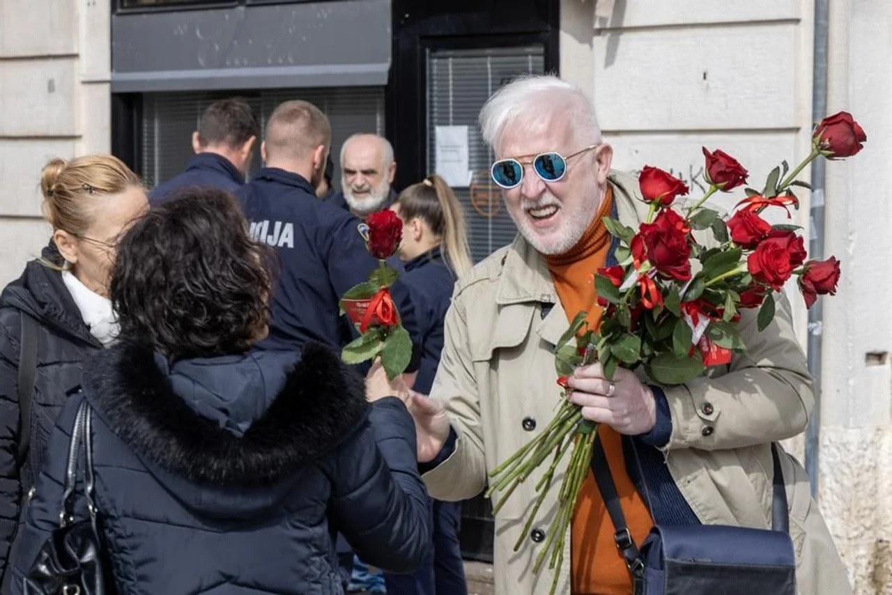 Battifiaca sa sunčanim naočalama nasmijan predaje crvenu ružu ženi na ulici.