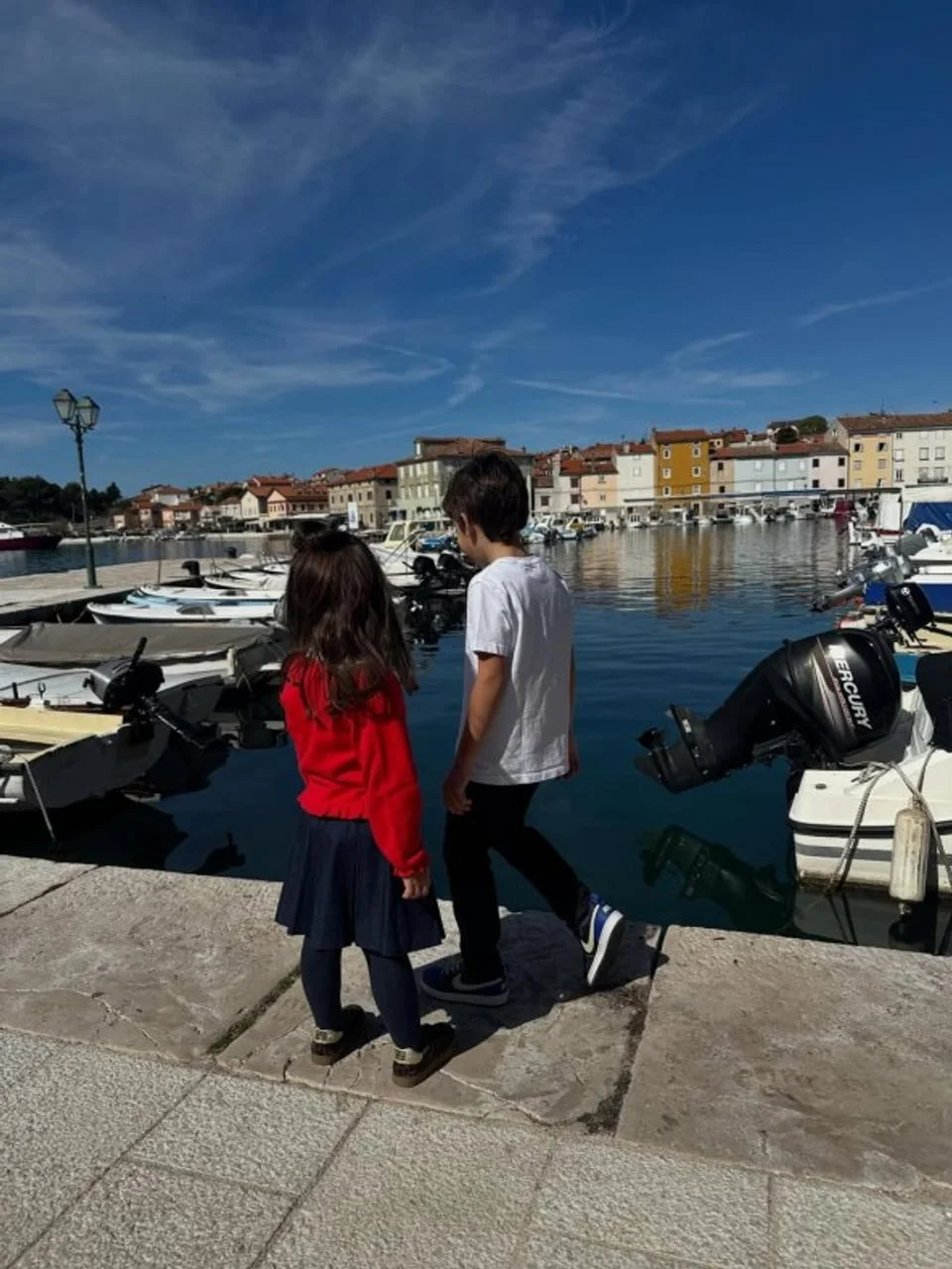 Dvoje male djece stoji na kamenoj rivi uz brodice u luci.