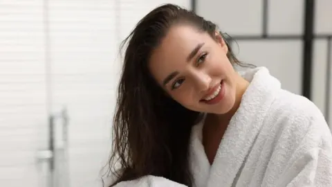 Nasmijana žena s mokrom kosom u bijelom kupaonskom ogrtaču.