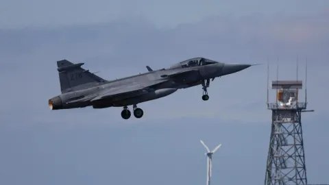 Sivi borbeni zrakoplov u letu iznad zračne baze s radarom u pozadini.