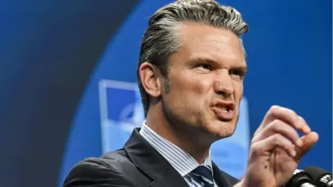 Pete Hegseth tijekom govora s ozbiljnim izrazom lica i podignutom rukom.