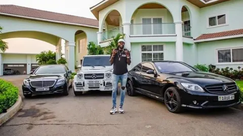 Emmanuel Adebayor stoji ispred luksuzne vile i prestižnih automobila na prilazu.