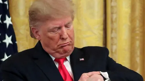 Donald Trump u tamnom odijelu s crvenom kravatom gleda na ručni sat.