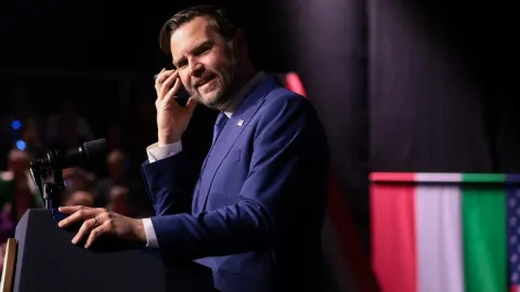 JD Vance u plavom odijelu drži mobitel na uhu dok stoji za govornicom.