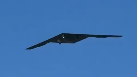 Crna stealth letjelica tipa letećeg krila u letu na čistom plavom nebu.