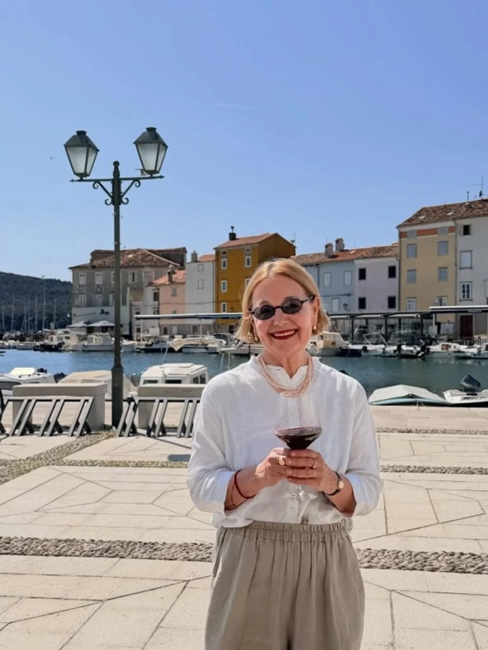 Vitomira Lončar s čašom vina ispred luke s brodovima i kamenim kućama.
