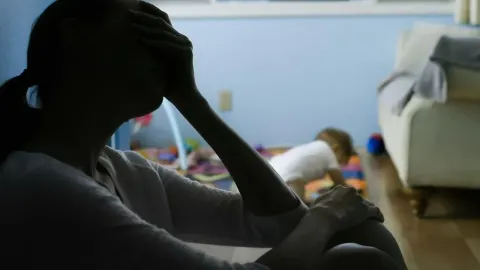 Majka drži ruku na čelu u zamračenoj sobi dok se dijete igra u pozadini.