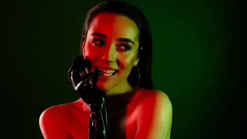 Portret nasmiješene žene pod crvenim i zelenim svjetlom dok grize crnu rukavicu.