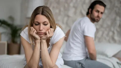 Zamišljena žena sjedi na rubu kreveta dok muškarac u pozadini gleda u stranu.