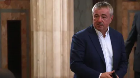 Dušan Bajatović u plavom sakou i bijeloj košulji stoji u unutrašnjem prostoru.