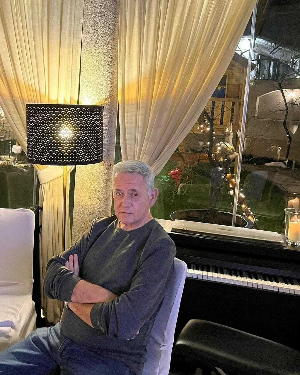 Stariji muškarac prekriženih ruku sjedi pored klavira u interijeru