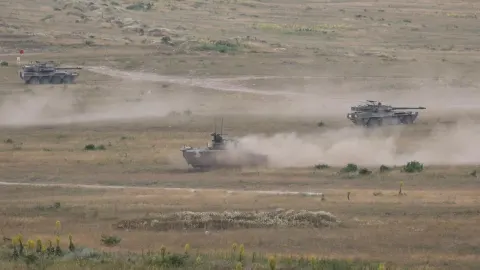 Tri oklopna vojna vozila u pokretu na pješčanom terenu podižu prašinu.