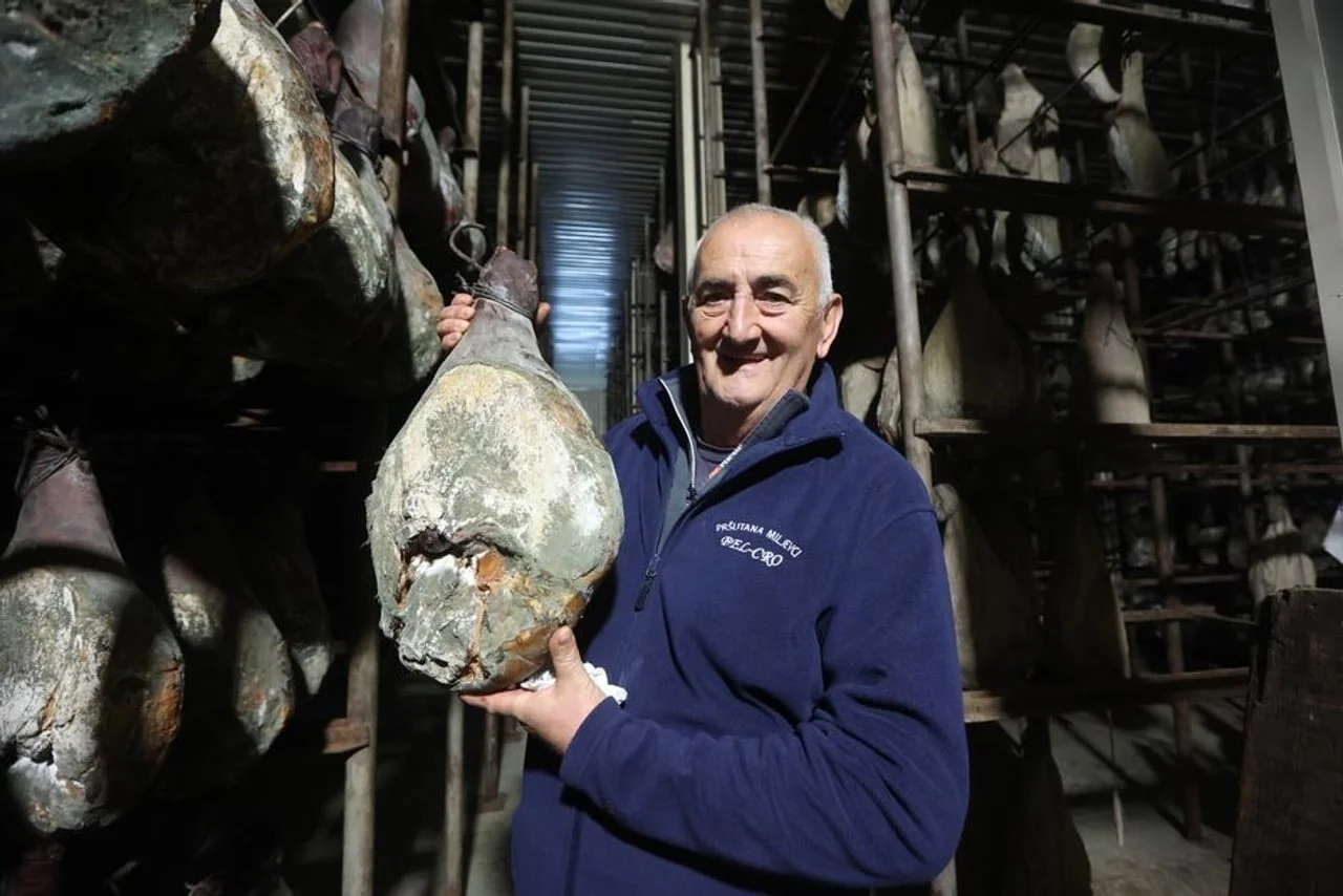 Nasmijani muškarac u pršutani drži cijeli drniški pršut ispred polica za zrenje.