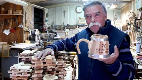 Majstor u radionici drži tradicionalni drveni susak pored hrpe drvenih daščica.