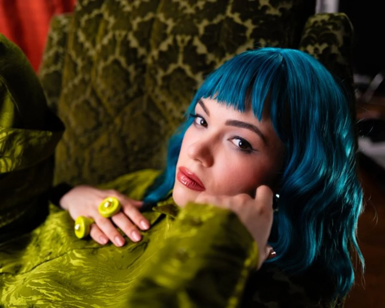 Gina Damjanović s plavom perikom leži na zelenom baršunastom naslonjaču