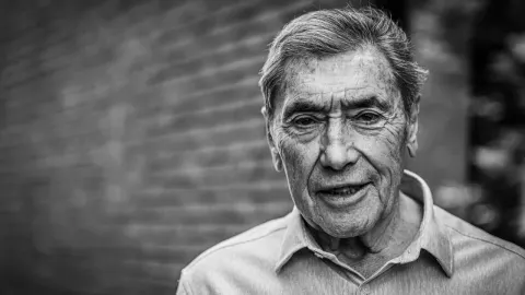 Crno-bijeli portret starijeg muškarca ozbiljnog izraza lica ispred zida od cigle.