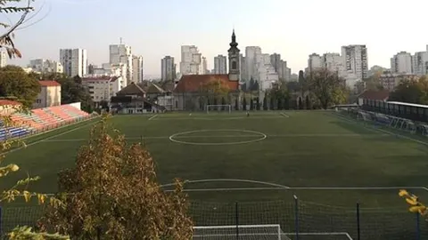 Nogometno igralište FK Bežanija s pogledom na crkvu i stambene zgrade u pozadini.