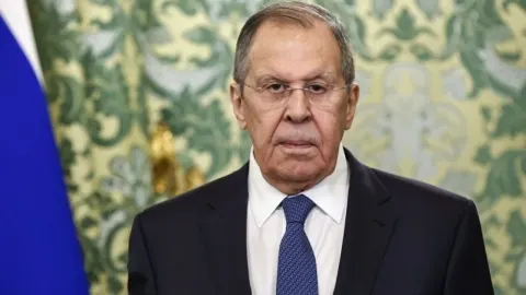 Sergej Lavrov u tamnom odijelu s plavom kravatom ispred ruske zastave i ukrasnih tapeta.
