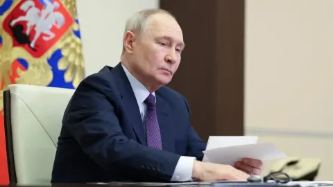 Vladimir Putin sjedi za radnim stolom ispred ruske zastave s dokumentima u ruci.