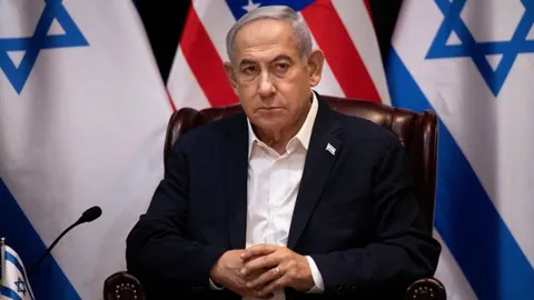Benjamin Netanyahu sjedi ispred zastava Izraela i SAD-a ozbiljnog izraza lica.