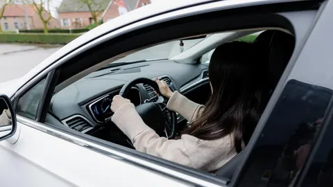 Žena sjedi za upravljačem parkiranog bijelog automobila ispred kuća.