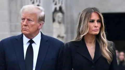 Donald i Melania Trump u ozbiljnom izdanju stoje jedno uz drugo ispred kamene građevine.