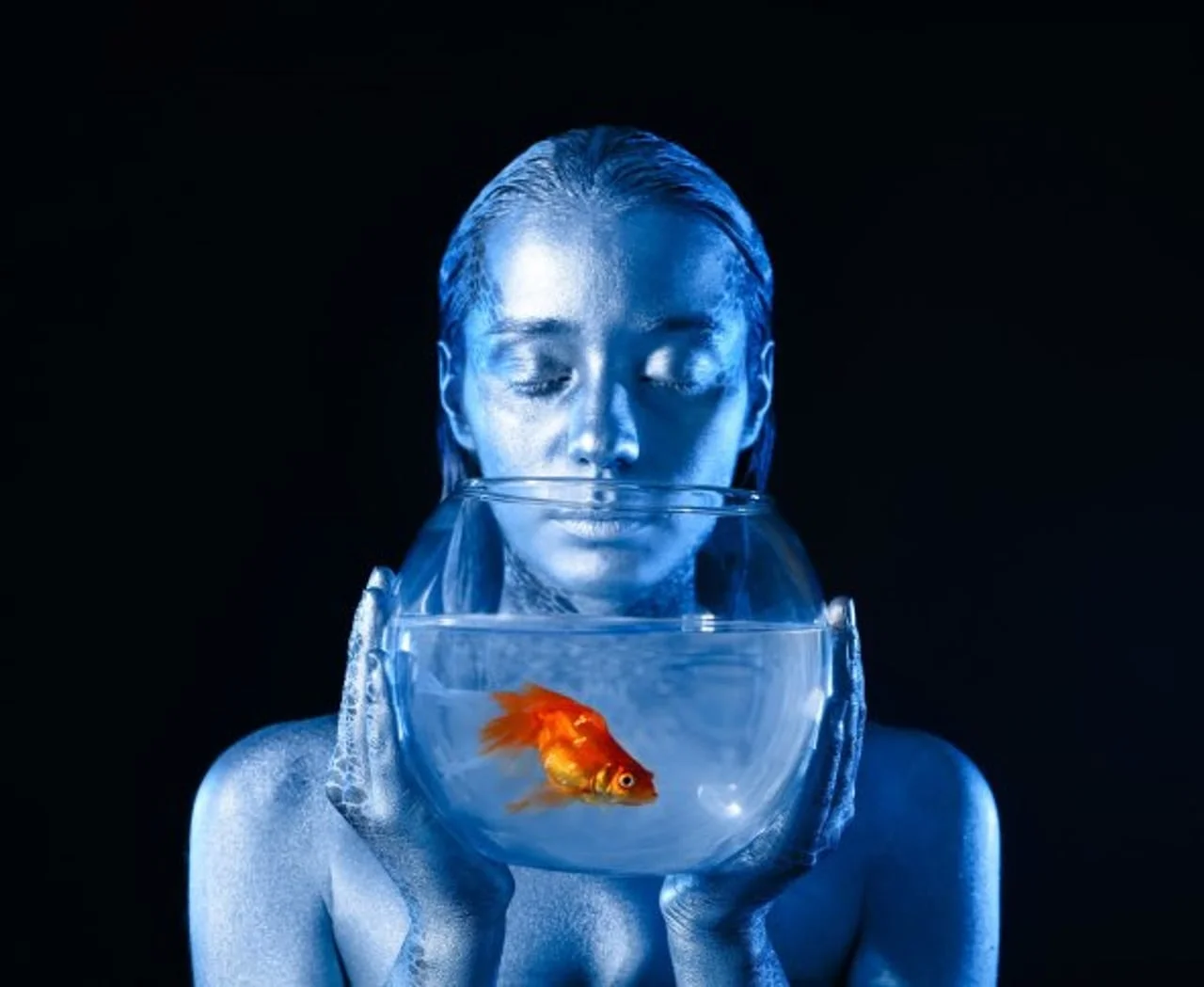 Žena plave kože drži akvarij sa zlatnom ribicom ispred lica.