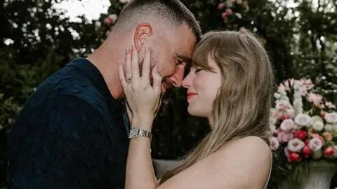 Taylor Swift i Travis Kelce u intimnom zagrljaju prislonjenih čela s cvijećem u pozadini.