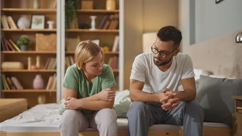 Mladi bračni par sjedi na rubu kreveta u spavaćoj sobi s ozbiljnim izrazima lica.