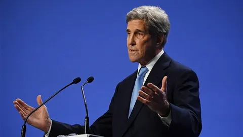 John Kerry u odijelu govori za govornicom s mikrofonima na plavoj pozadini.