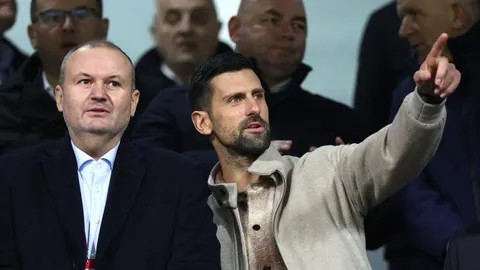 Novak Đoković u bež jakni pokazuje rukom prema naprijed u gledalištu.