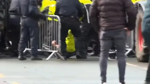 Policajci u crnim uniformama i prosvjednici iza metalne ograde na cesti.