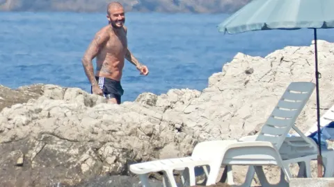 David Beckham bez majice na stjenovitoj plaži pokazuje tetovaže i mišiće.