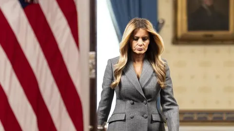 Melania Trump u sivom sakou stoji ispred američke zastave u Bijeloj kući.