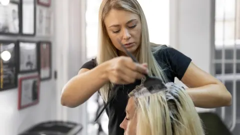 Plavokosa frizerka u crnoj majici nanosi boju kistom na kosu klijentice u salonu.