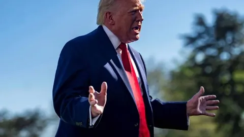 Donald Trump u tamnom odijelu s crvenom kravatom govori s raširenim rukama vani.