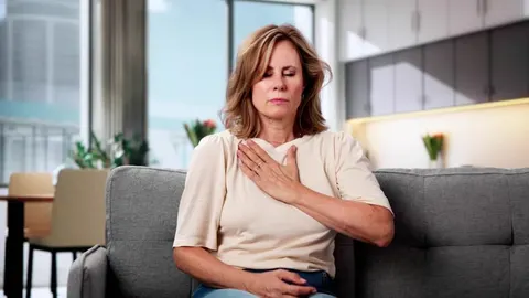 Žena se drži za prsa s izrazom nelagode dok sjedi na kauču u dnevnom boravku.
