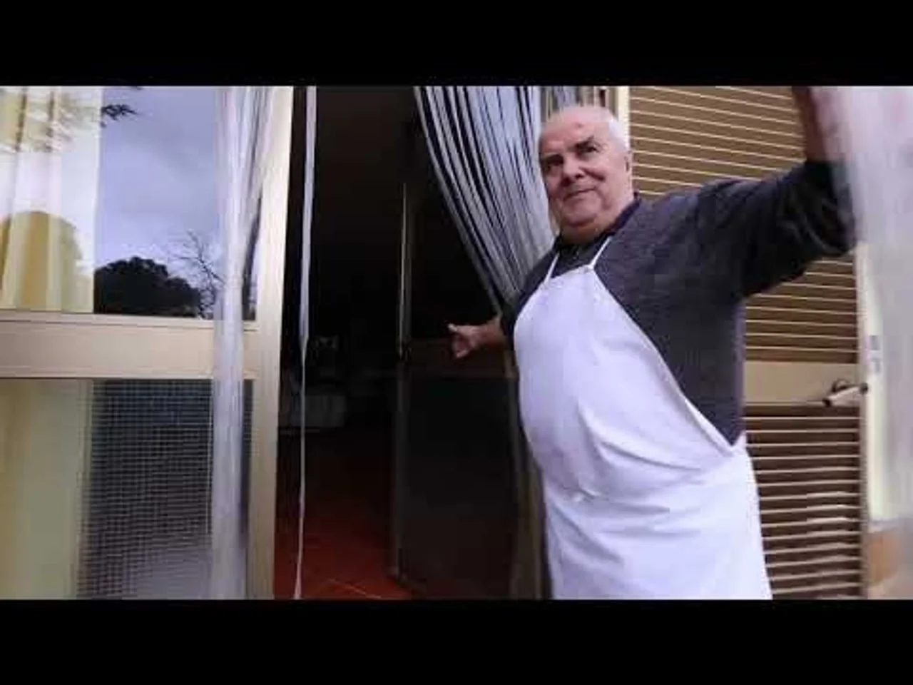 Stariji muškarac s bijelom pregačom stoji na ulazu u kuću.