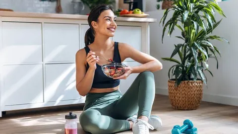 Mlada žena u sportskoj odjeći sjedi na podu kuhinje i jede zdravi doručak.