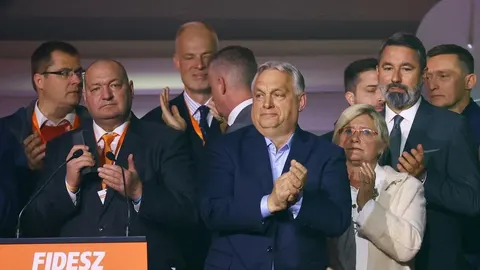 Viktor Orbán stoji na pozornici okružen suradnicima ispred znaka stranke Fidesz.