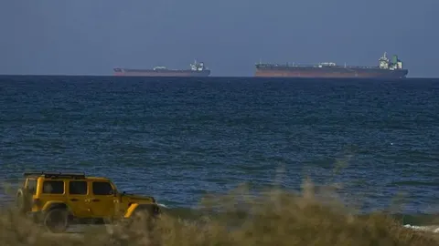 Naftni tankeri na pučini i žuto terensko vozilo na obali.